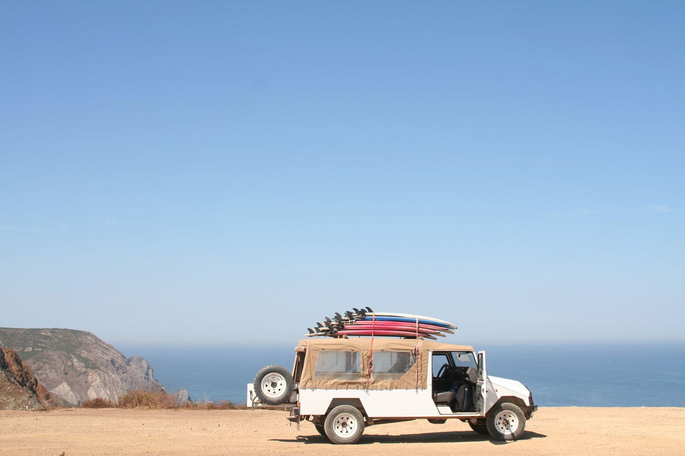 coche blanco descapotable con cinco tablas de surf de diferentes colores en la baca del coche. El coche está sin pasajeros en la arena y de fondo el mar